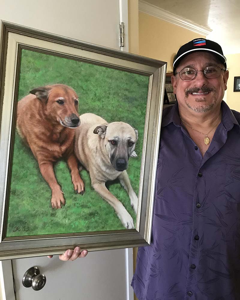 Man holding framed painting of two dogs lying on grass.