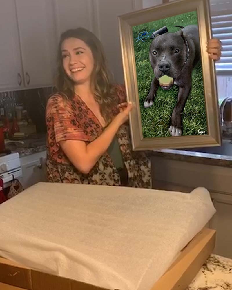 Woman holding framed pet portrait of a pit bull holding a tennis ball.