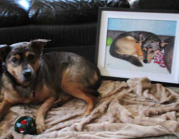 Dog lying on a blanket next to a framed painting of a dog.