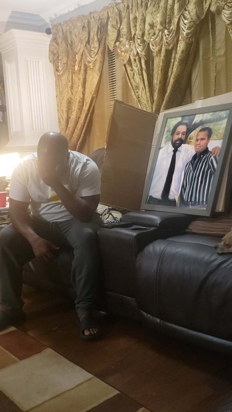Man sitting on couch, head in hands, next to framed portrait of two men.