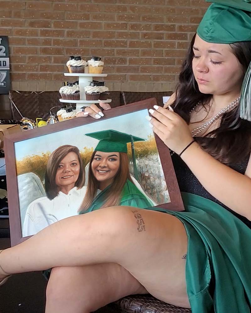 Graduate in cap and gown holding framed portrait of herself and another woman.