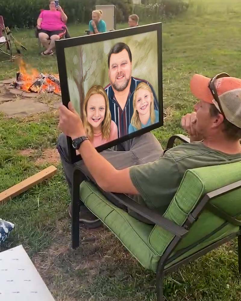 Man in a green chair holding a framed painting of a man and two girls in front of a campfire.